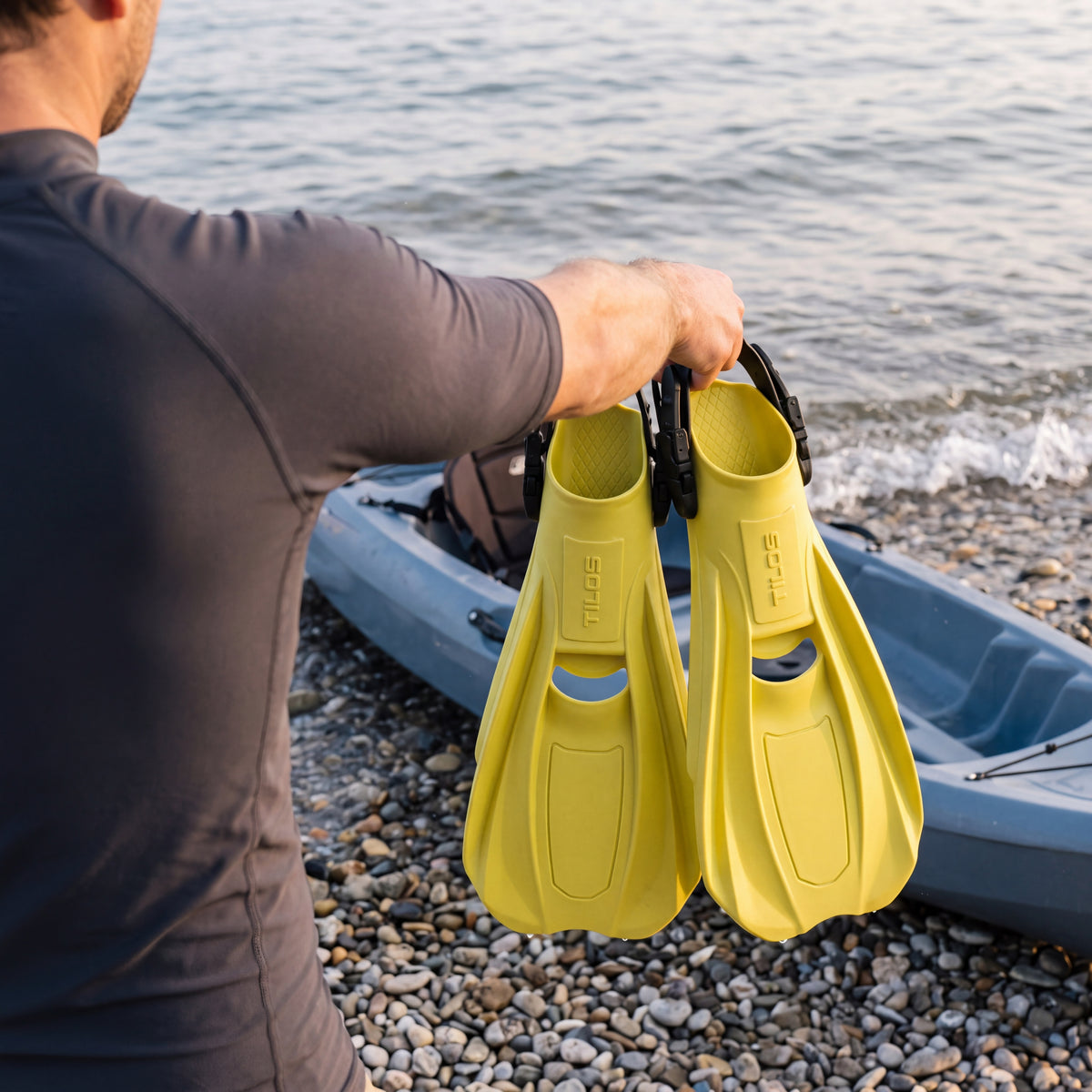 Lifestyle photo of a person carrying yellow Tilos Technix open heel travel fins by the straps beside a kayak on a pebble beach
