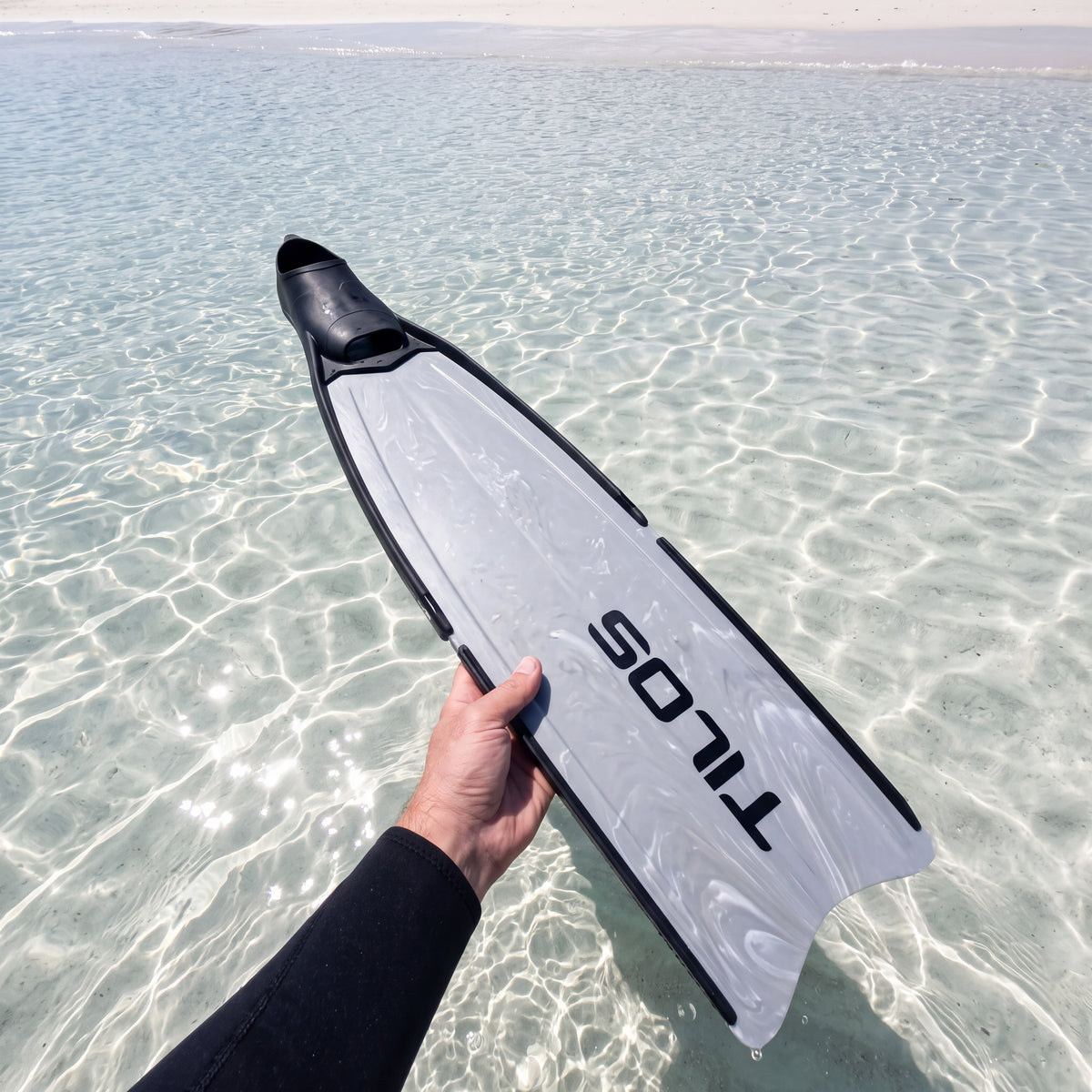 Freediver holding a Tilos Syryn full foot freediving fin in Black Camo above clear shallow water, first person view of the long marbled blade