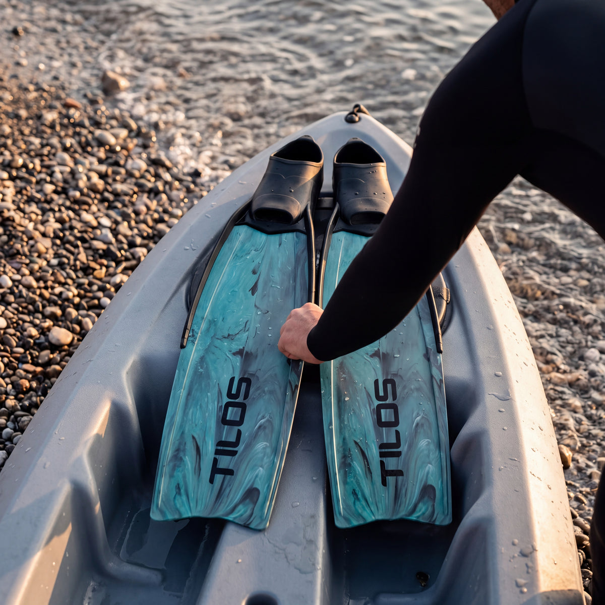 Freediver laying a pair of Tilos Syryn full foot freediving fins in Green Camo across a kayak at a pebbly shoreline