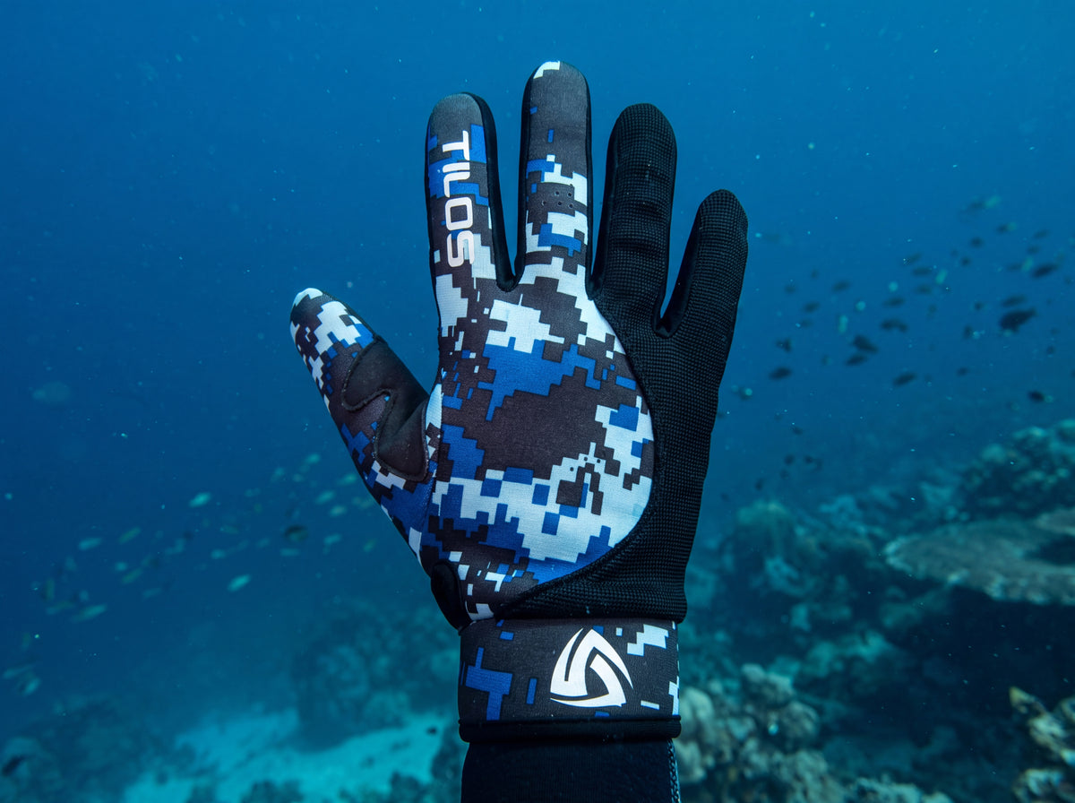 A scuba diver raises a gloved hand underwater wearing Digital Blue Tilos 1.5mm Tropical-X Mesh gloves, the blue pixel camo back panel and white TILOS wordmark square to the camera.