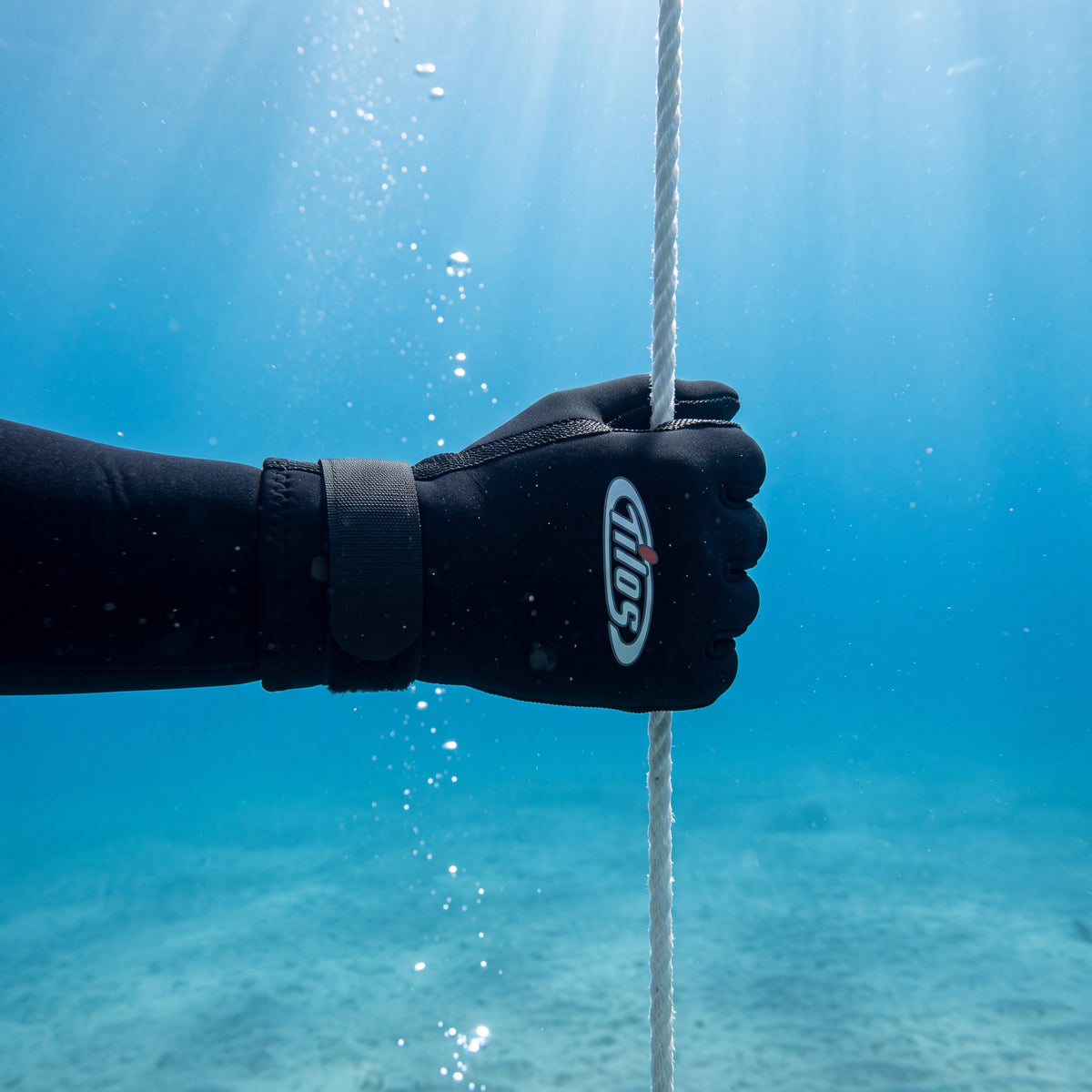 Underwater view of a diver wearing a black Tilos Supratex dive glove gripping a white descent line in clear blue water