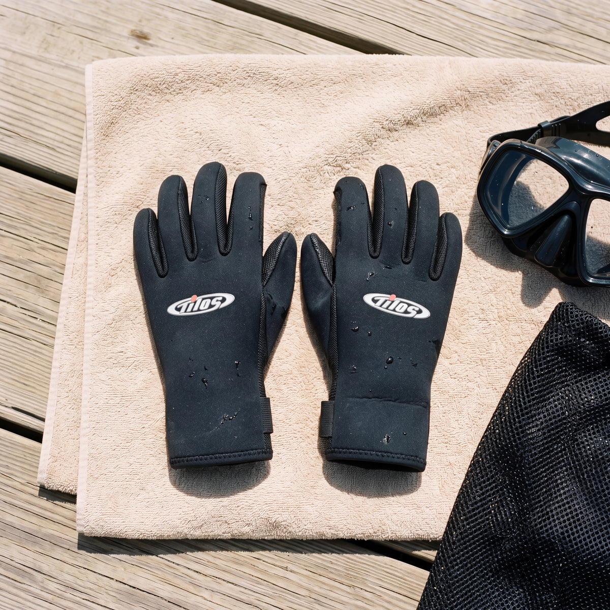 Lifestyle flat lay of a pair of black Tilos Supratex dive gloves on a beach towel on a wooden dock beside a dive mask and mesh gear bag