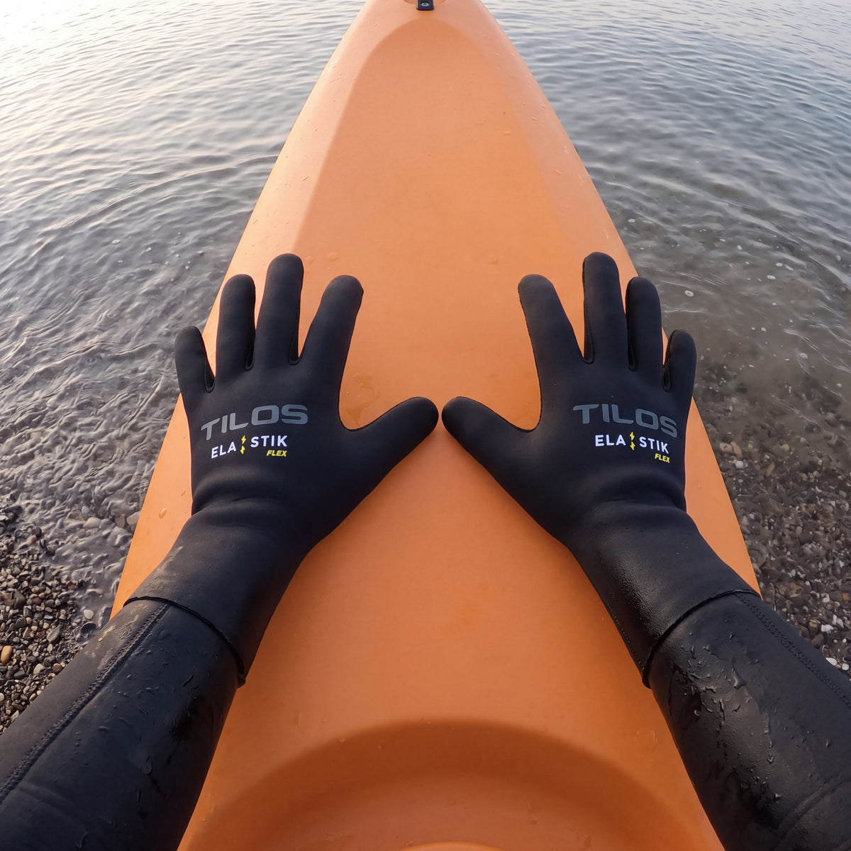 First person view of a kayaker wearing black Tilos 2mm Elastik SuperStretch gloves, both hands pressed on the deck of an orange sit-on-top kayak at a pebble shoreline