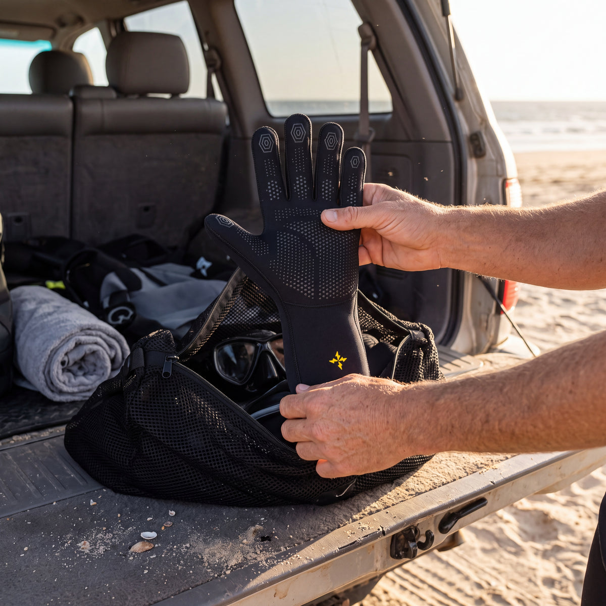 Diver holding up a black Tilos 2mm Elastik SuperStretch glove palm side out, showing the hexagon fingertip grip and dotted palm, before packing it into a mesh dive bag at the back of a car by the beach