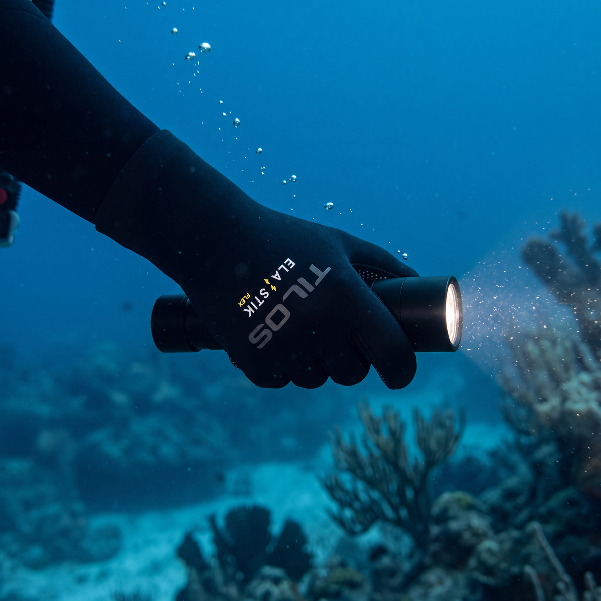 Diver underwater wearing a black Tilos 2mm Elastik SuperStretch glove holding a dive torch over a coral reef