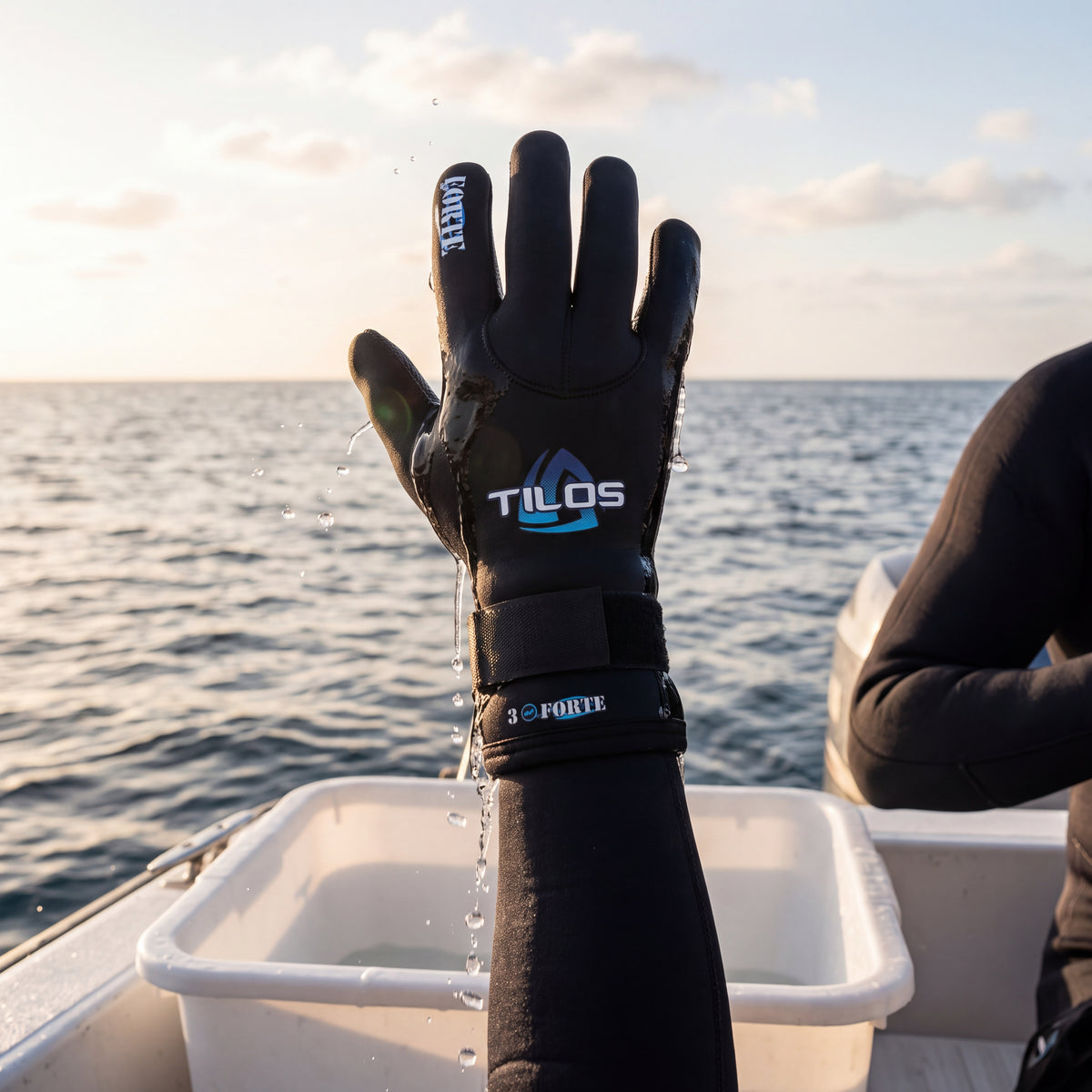 Diver on a boat after a dive wearing a black Tilos 3mm Forte Velcro Glove, lifting a hand out of a freshwater rinse tub at sunset