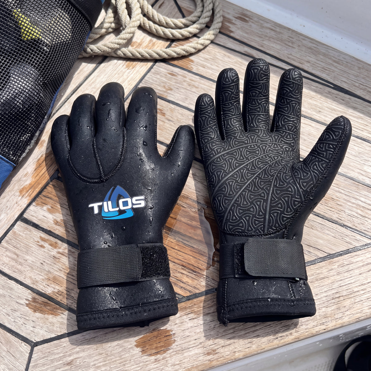Lifestyle flat lay of a pair of black Tilos 5mm Titanium neoprene dive gloves on a wet boat deck, one showing the logo and one the grip palm.