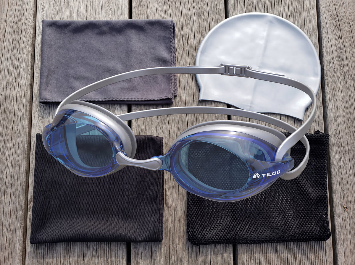 Top down lifestyle flat lay of the Tilos Vynix swimming goggles in blue with silver trim on wooden decking beside a quick dry towel, swim cap and mesh gear bag