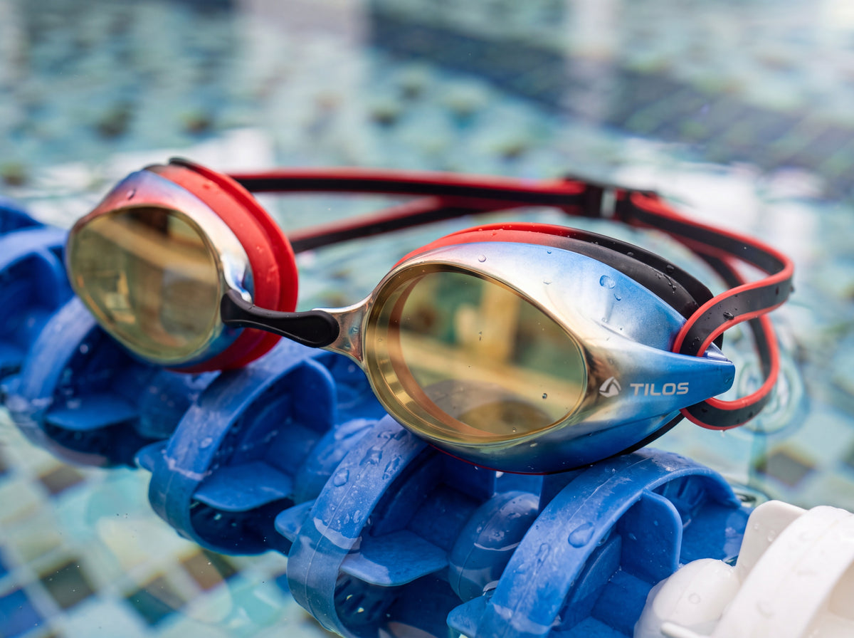 Poolside lifestyle close detail of the Tilos Vynix swimming goggles in red and black with gold mirrored lenses resting on a lane rope