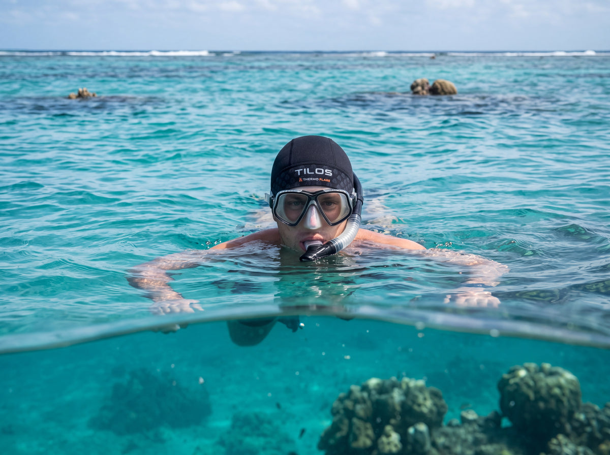 A snorkeler floats at the surface over a shallow reef, photographed from water level with the waterline across the frame, wearing a black Tilos Thermoflare neoprene skull cap.