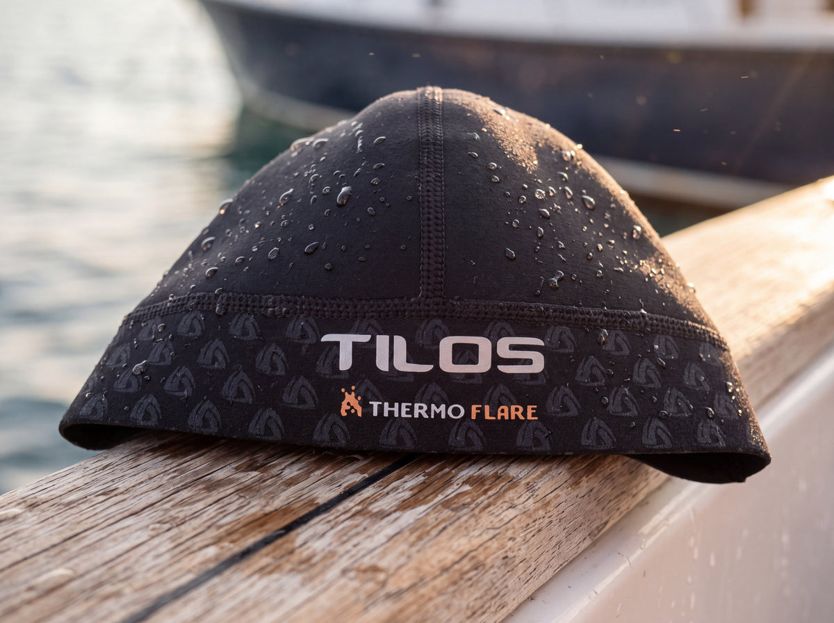 Lifestyle detail of a freshly rinsed black Tilos Thermoflare neoprene skull cap drying on a teak boat rail after a dive, water beaded on the neoprene, the silver TILOS wordmark and copper THERMO FLARE mark sharp on the front band.