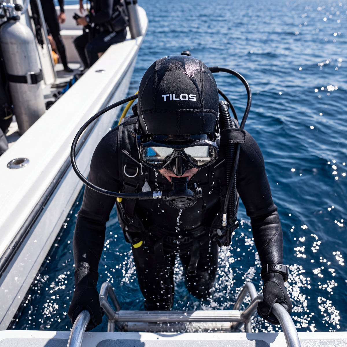 A diver wearing a black Tilos 3mm neoprene dive hood climbs a boat ladder out of the ocean, seawater running off the hood and shoulders.