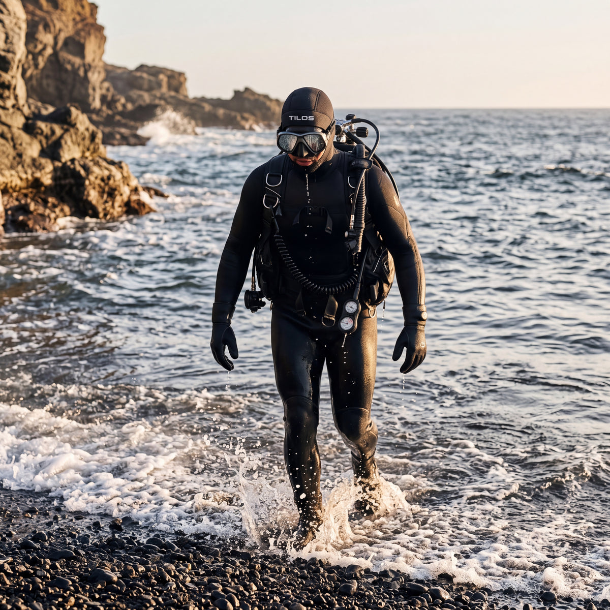 A diver wearing a black Tilos 3mm neoprene dive hood walks out of the surf onto a rocky shore at sunset.