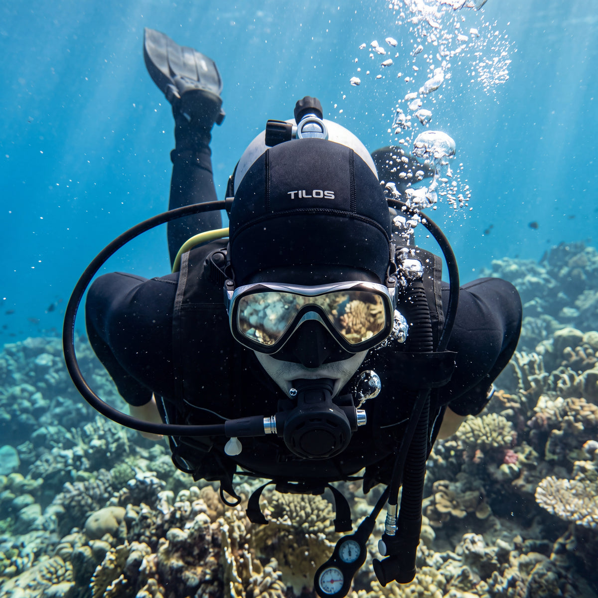 A diver wearing a black Tilos 3mm neoprene dive hood swims over a coral reef underwater.