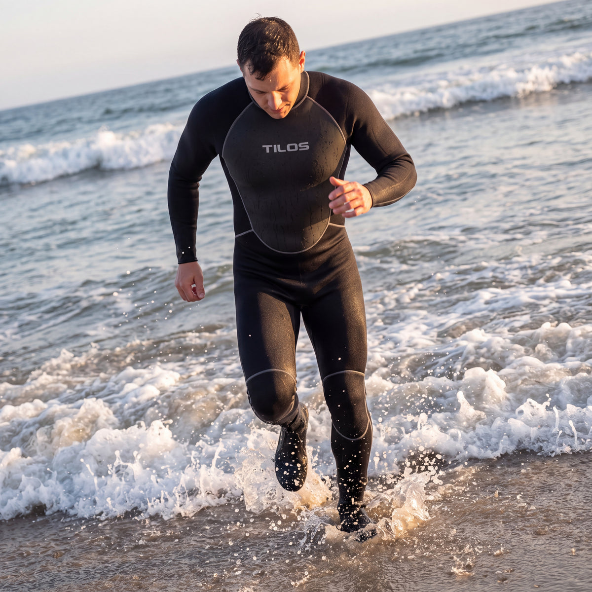 Diver striding out of breaking surf at golden hour wearing a black Tilos J3204BK full wetsuit, silver Tilos chest wordmark visible.