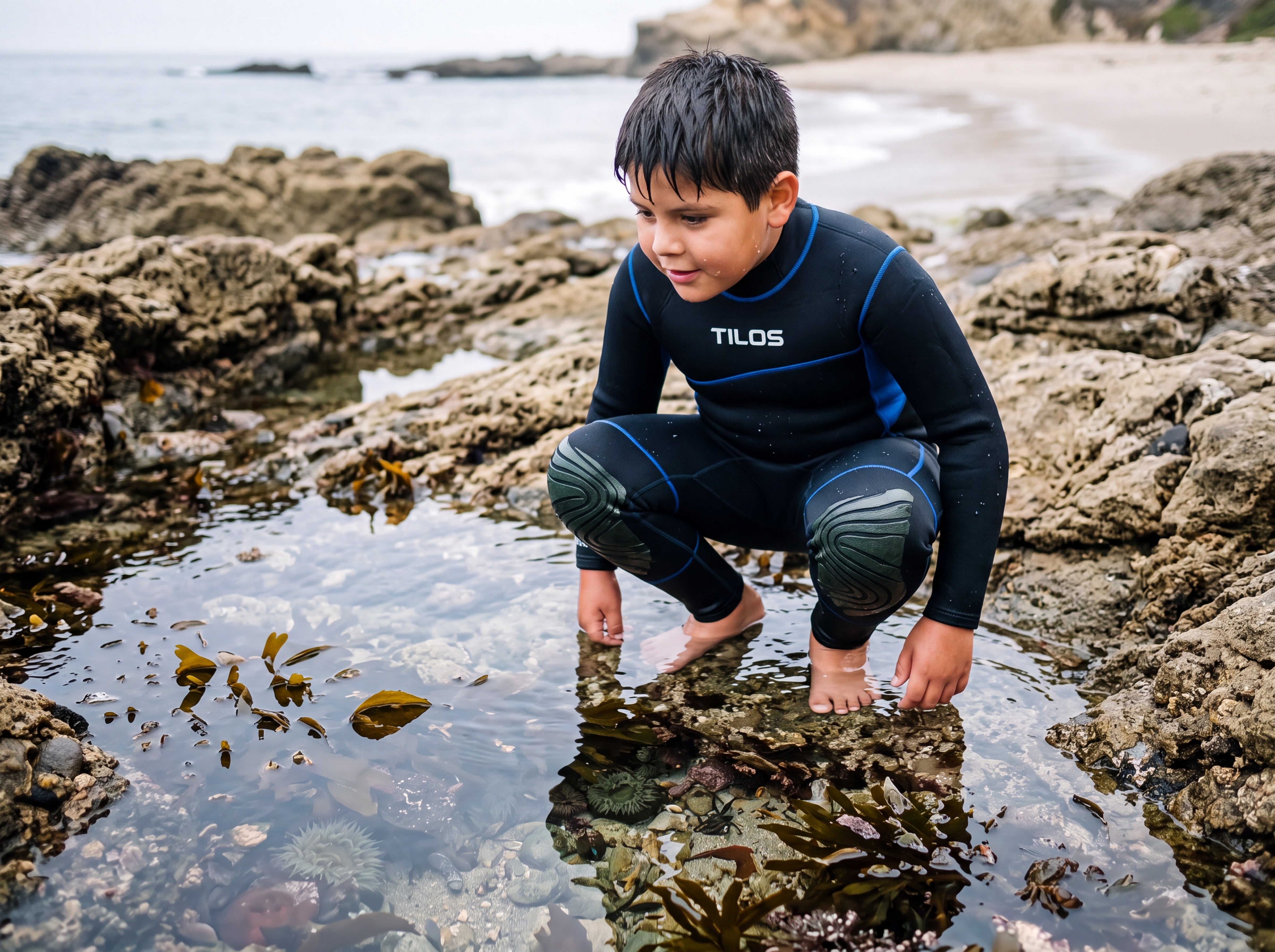 A child wearing the Tilos Kid's 3/2mm Adventure Steamer wetsuit while crouching to explore a rocky coastal tidepool