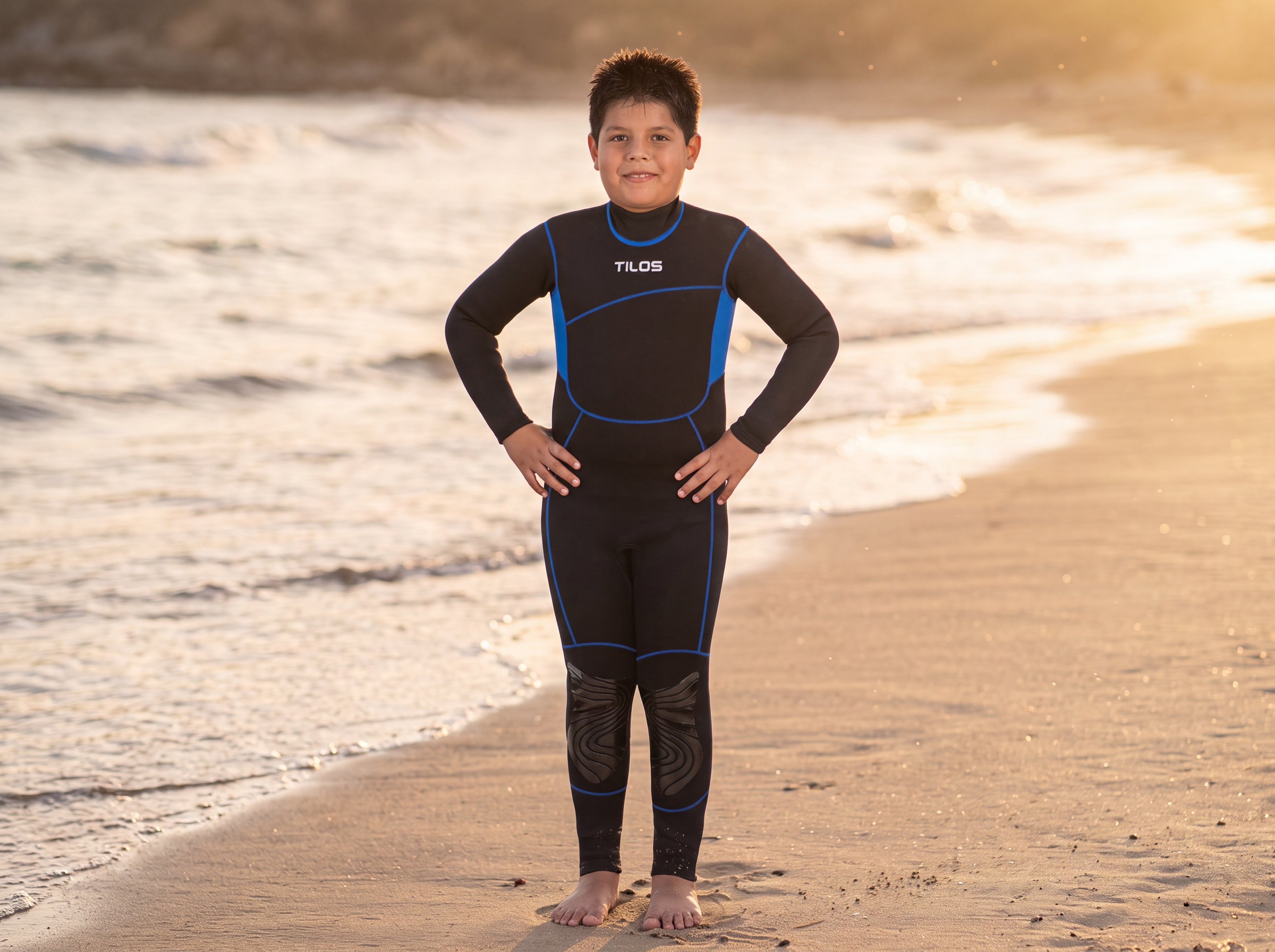 A child wearing the Tilos Kid's 3/2mm Adventure Steamer wetsuit standing on the shoreline at golden hour, ready to swim