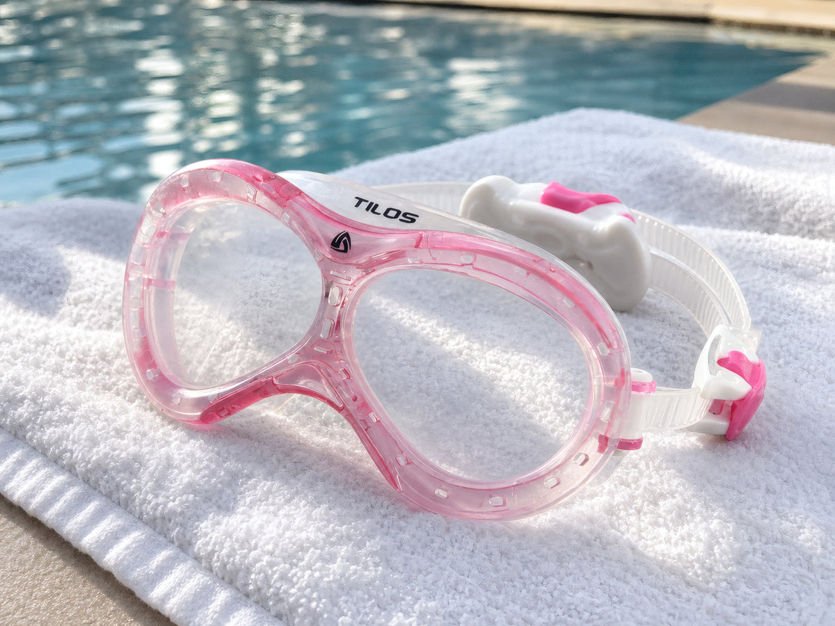 Pink Tilos Dyno toddler swim mask resting on a folded white towel poolside beside sunlit blue pool water