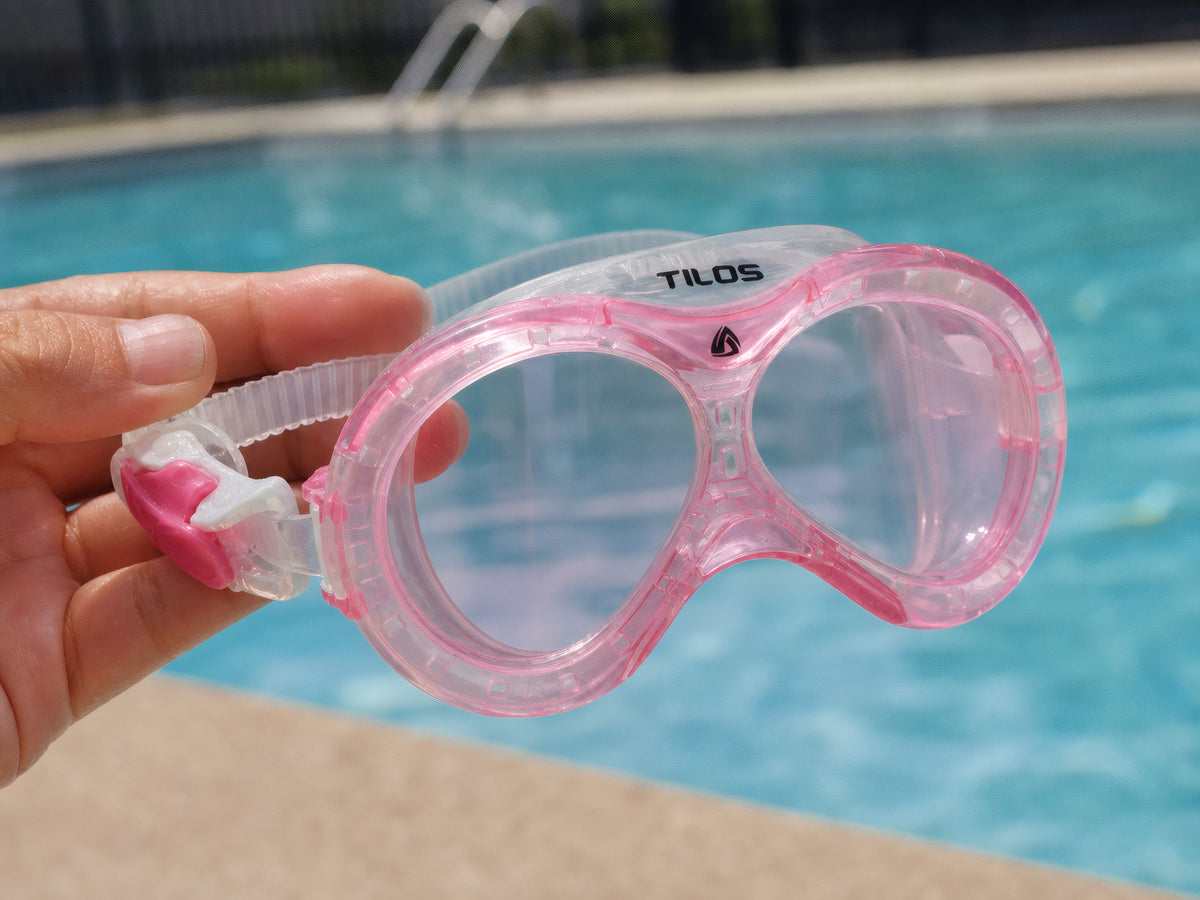 Hand holding the pink Tilos Dyno toddler swim mask poolside above bright blue swimming pool water