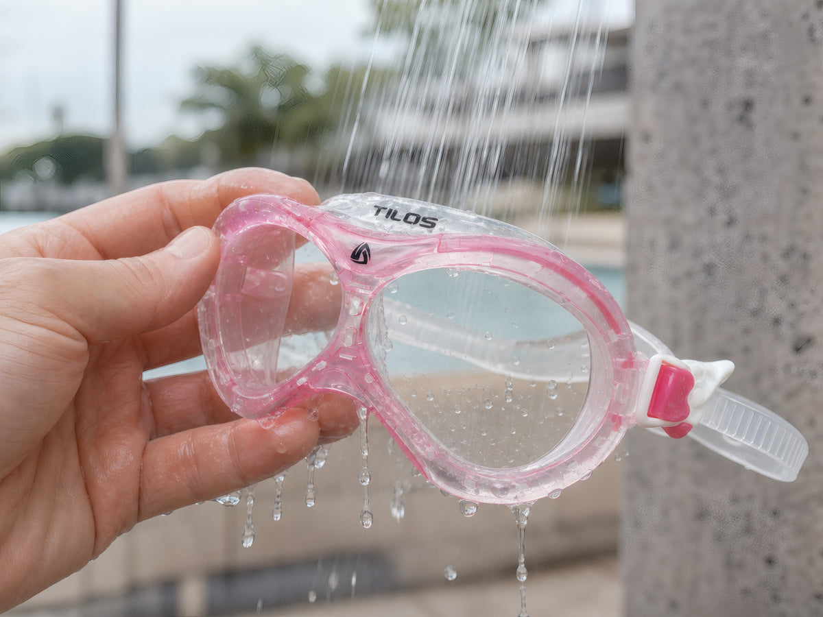 Hand rinsing the pink Tilos Dyno toddler swim mask under fresh running water at a poolside rinse shower
