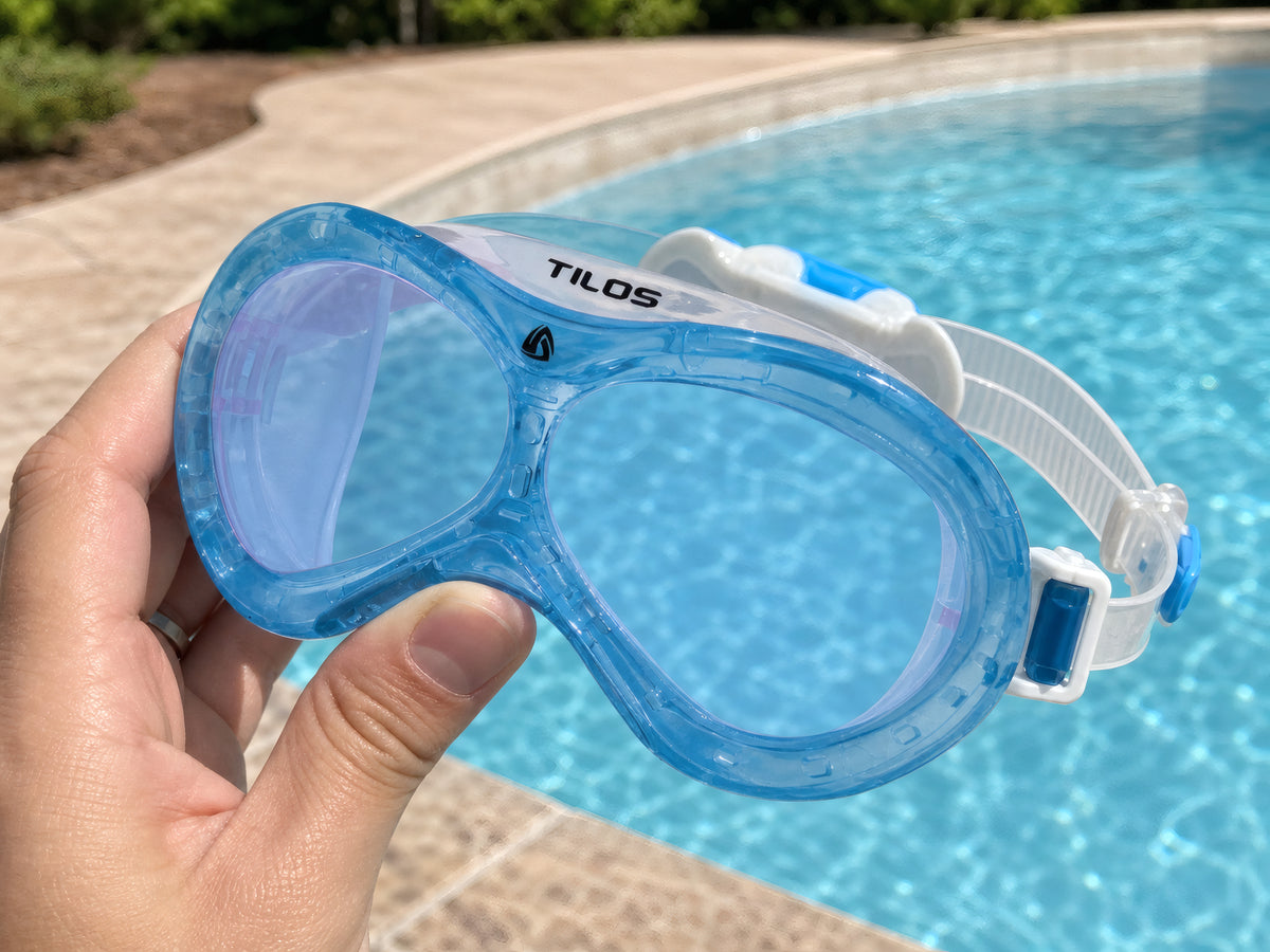 Hand holding the Transparent Blue Tilos Dyno toddler swim mask poolside above a curved swimming pool