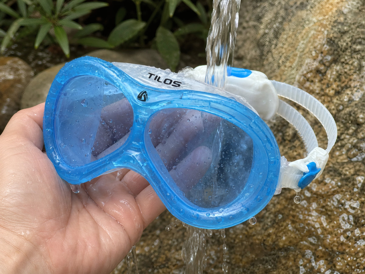 Lifestyle photo of a hand rinsing the Transparent Blue Tilos Dyno toddler swim mask under running water after swimming