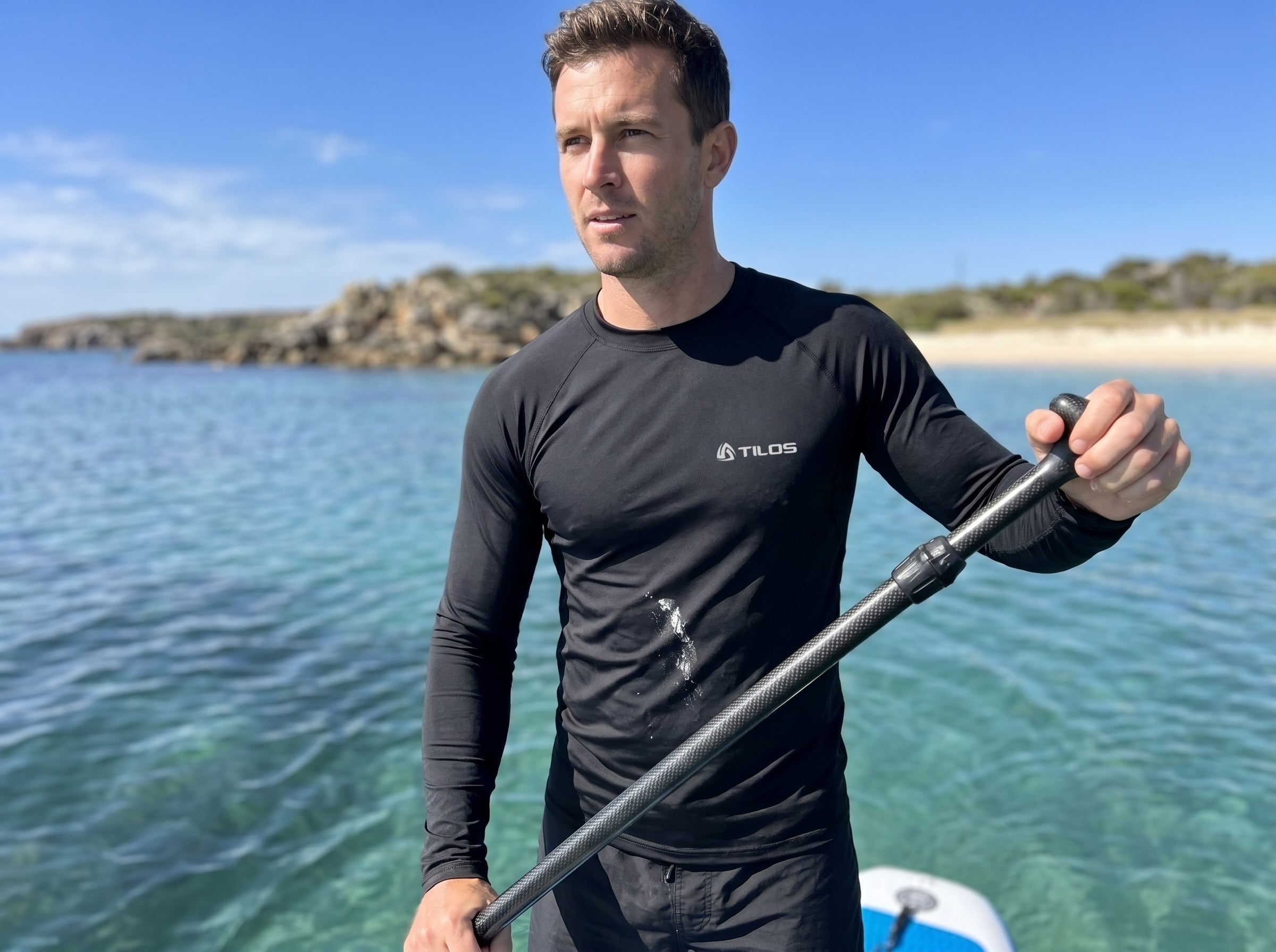 Man paddleboarding on calm ocean water wearing the Tilos Men's 6oz Anti-UV Long Sleeve Rash Guard in black