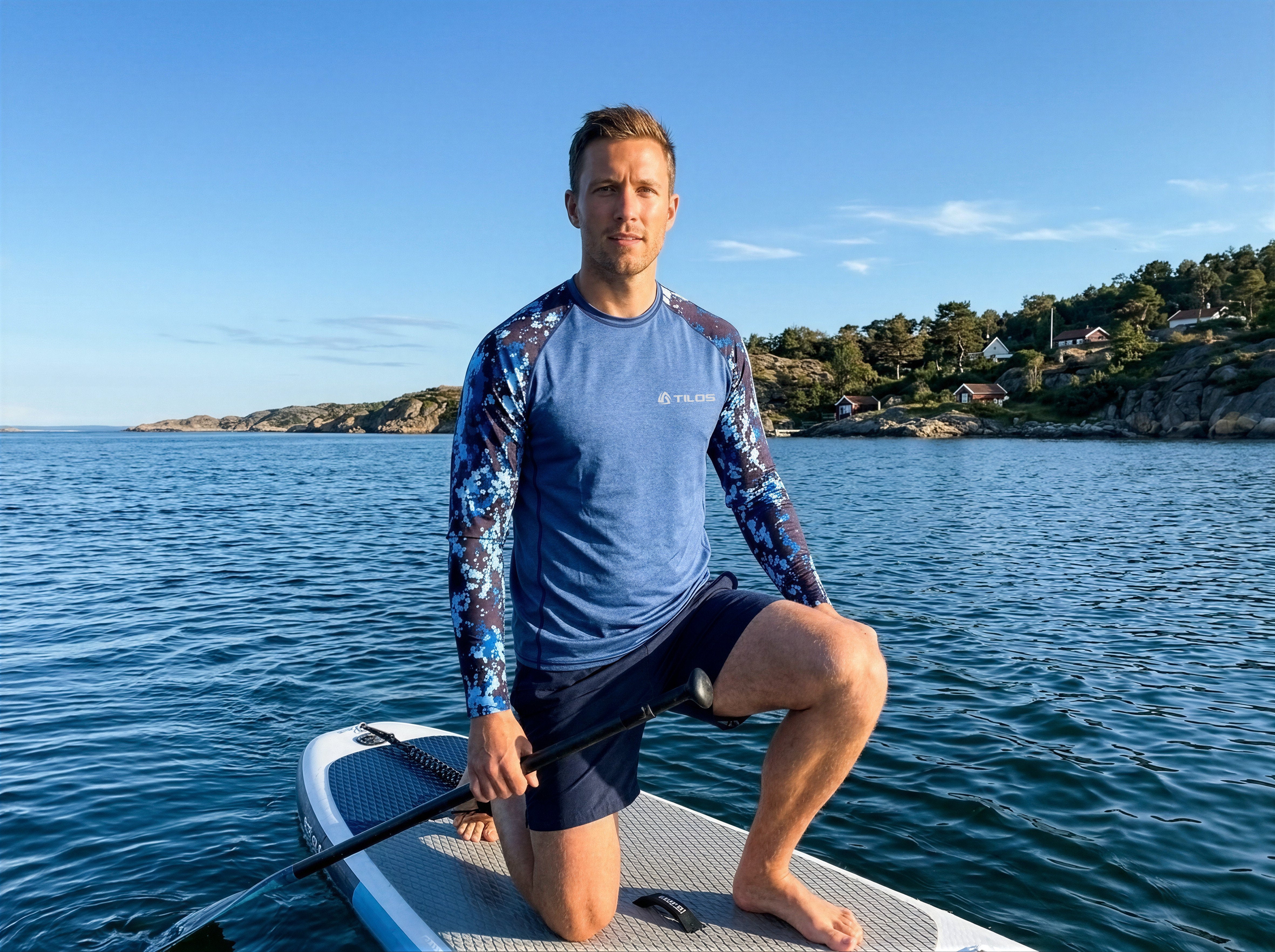Man kneeling on a paddleboard wearing the Tilos Men's 6oz Anti-UV Long Sleeve Rash Guard in Digital Blue camo