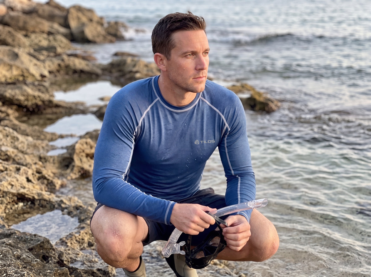 Man crouching at a rocky shore holding a dive mask wearing the Tilos Men's 6oz Long Sleeve Rash Guard in Heather Marine blue