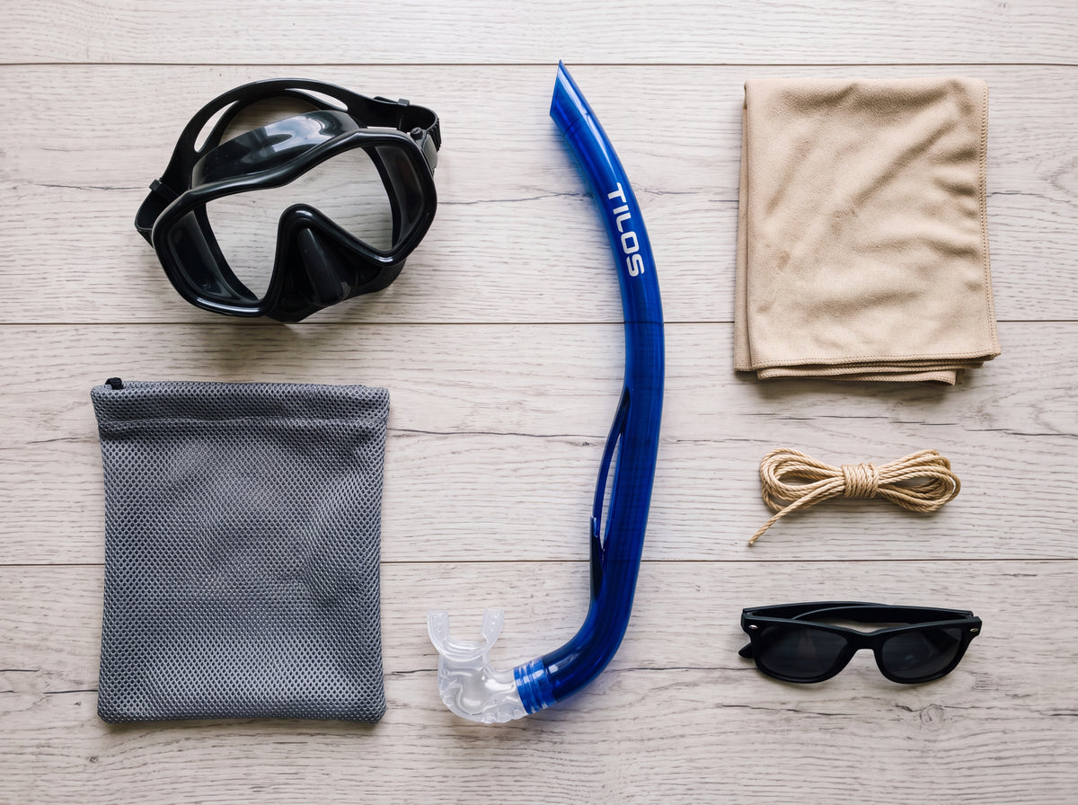 Top down lifestyle flat lay of the Tilos Basic J Snorkel in transparent blue packed beside a dive mask, quick dry towel and mesh gear bag