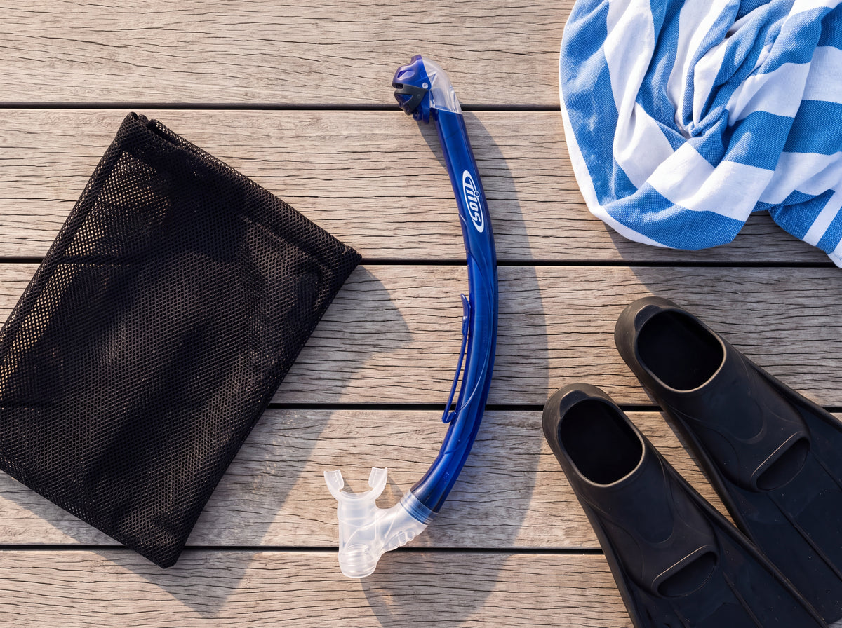 Top down lifestyle flat lay of the Tilos Oracle Dry Snorkel in transparent blue packed on a deck beside a mesh gear bag, towel and fins