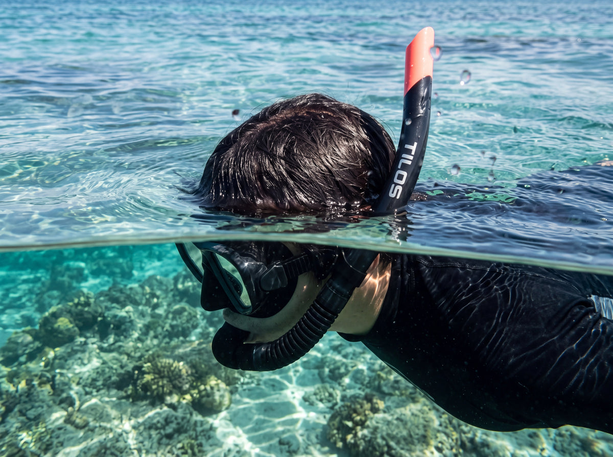 Split level view of a snorkeler floating face down at the surface of clear turquoise water, wearing a Black Silicone Tilos JX2 Flex Snorkel above a coral reef