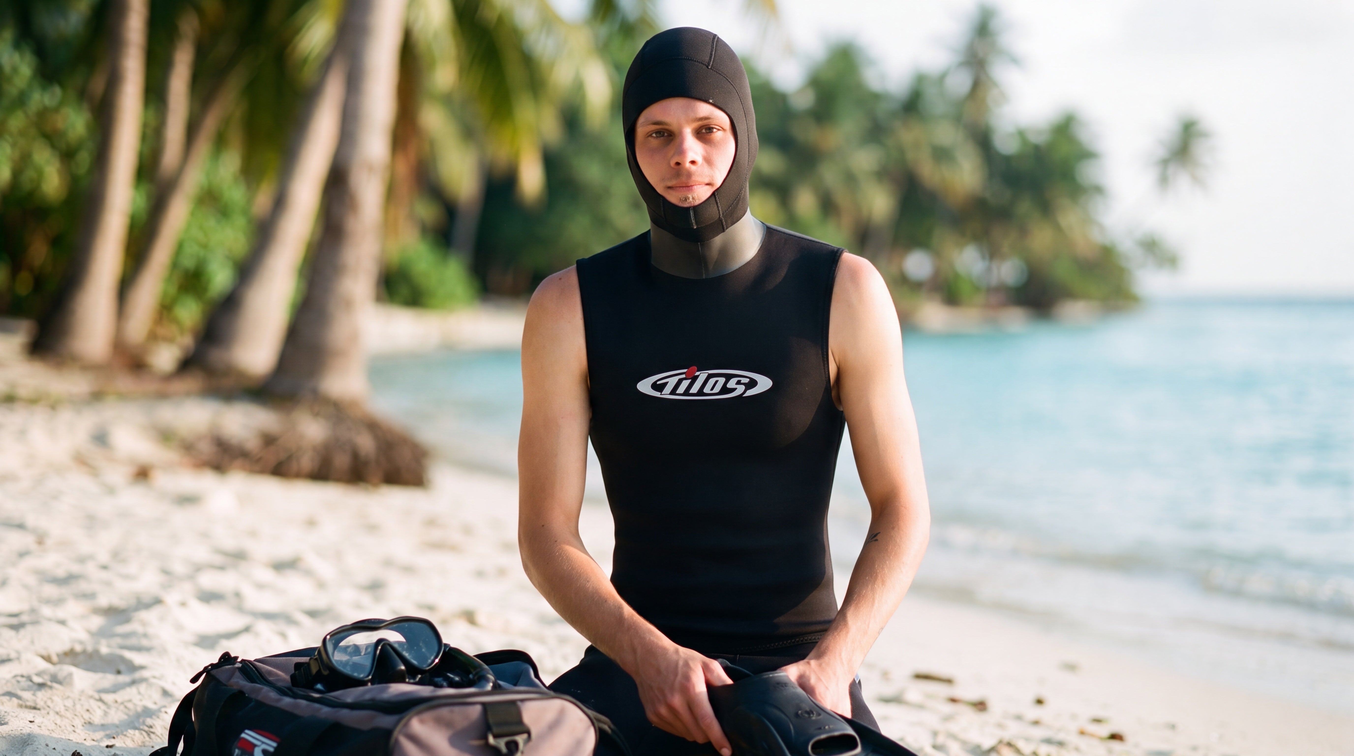 Diver kneeling on a palm-lined beach checking gear in the Tilos 5/3mm Superstretch Hooded Vest